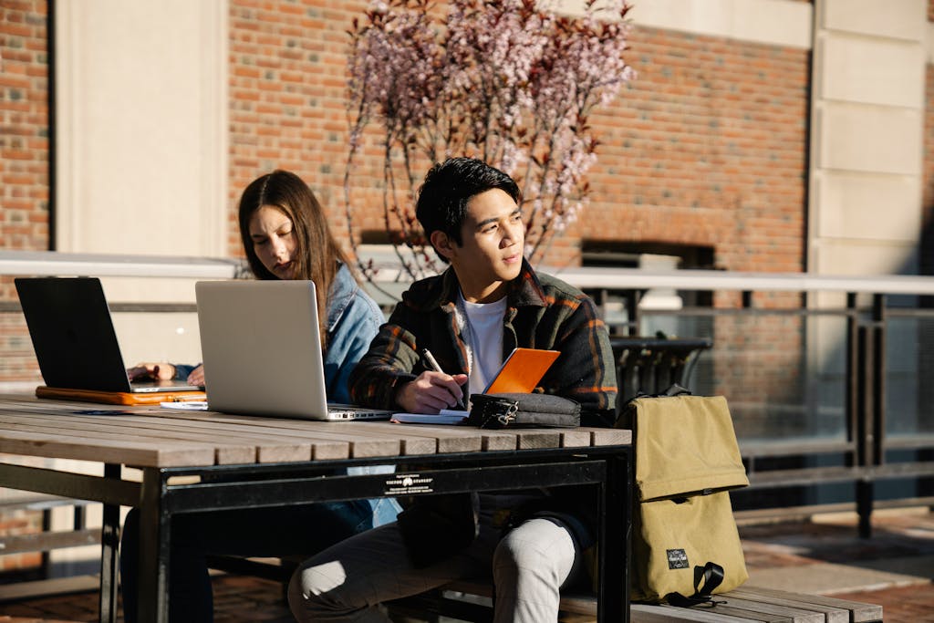 Two students studying outside on a sunny day with laptops and notebooks. Ideal for education and lifestyle themes.